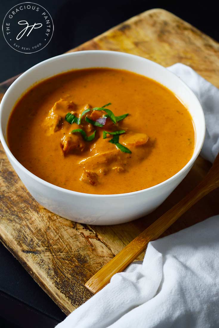 A white bowl sits filled with butter chicken. A wooden spoon and cloth napkin lay to the side.