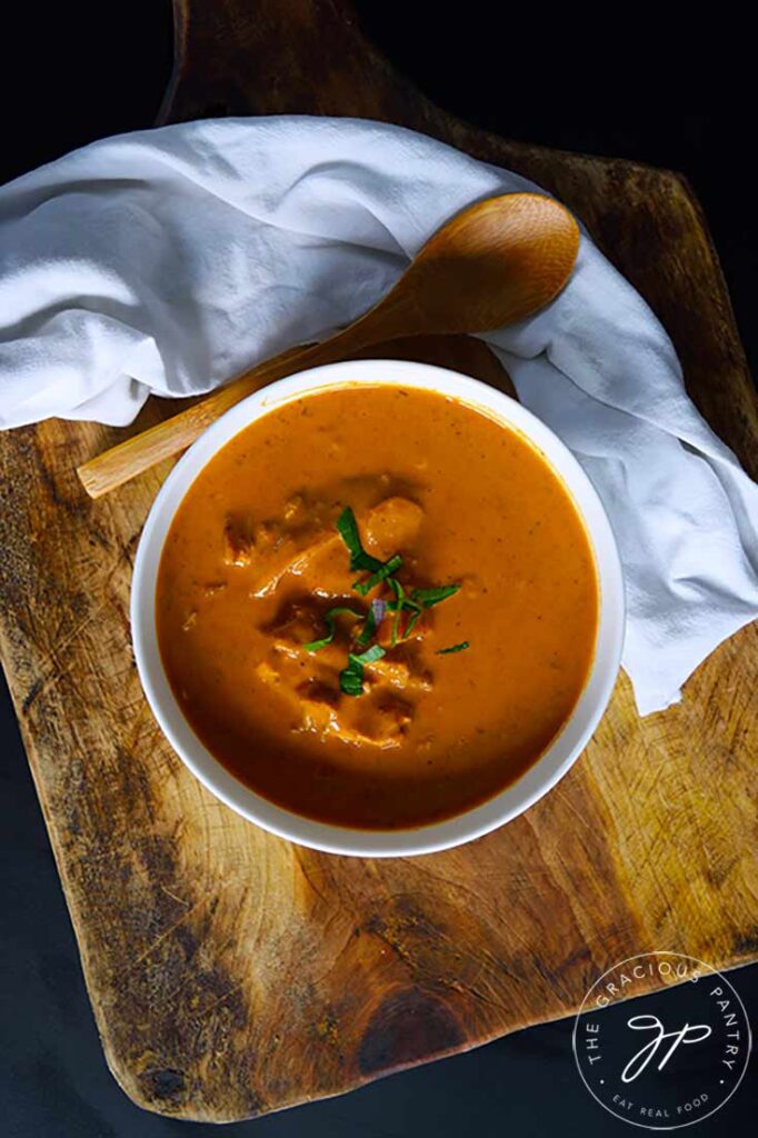 An overhead view looking down into a white bowl filled with butter chicken. A wooden spoon and napkin lay to the side.