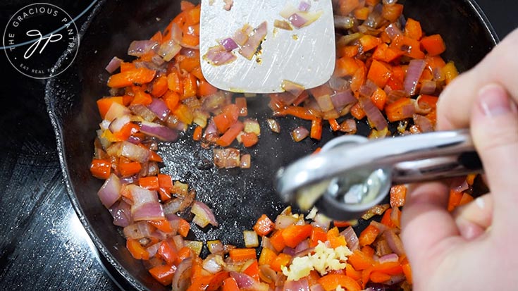 Pressing garlic into a skillet of chopped red peppers and onions.