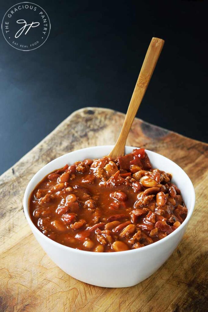 A white bowl holds a wooden spoon and is filled to the top with this Slow Cooker Cowboy Beans Recipe.