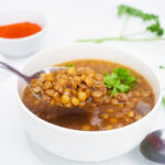 A spoon lifts some German Lentil Soup out of a white bowl. A small bowl of paprika sits on the table behind it.