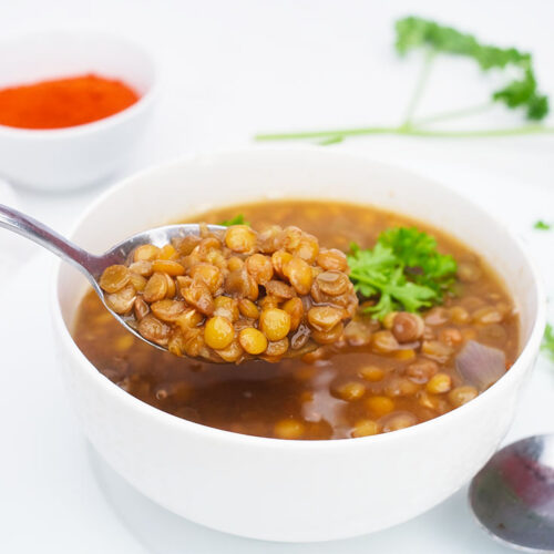 A spoon lifts some German Lentil Soup out of a white bowl. A small bowl of paprika sits on the table behind it.