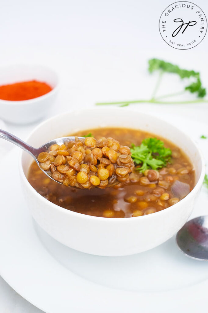 A spoon lifts some German Lentil Soup out of a white bowl. A small bowl of paprika sits on the table behind it.