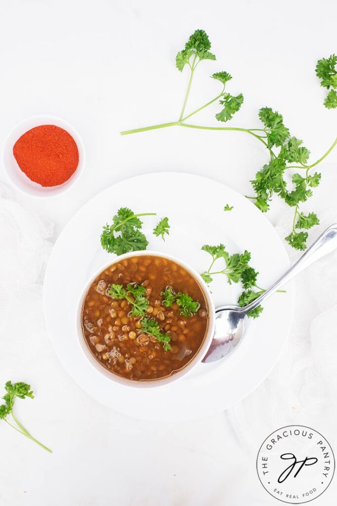 A white bowl filled with German Lentil Soup sits on a white plate and table. Fresh parsley is sprinkled around it.