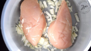 Two chicken breasts laying on top of chopped onions in a slow cooker.