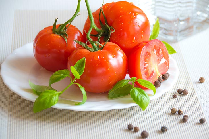 This list of superfoods includes tomatoes. This photo shows a white plate holds three whole tomatoes and one half of a cut tomato.