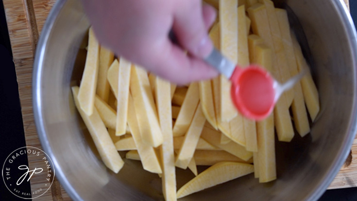 Adding oil to raw, cut, rutabaga fries in a mixing bowl.