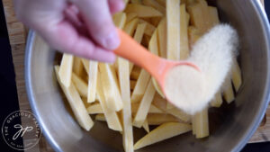 Adding garlic powder to raw, cut, rutabaga fries in a mixing bowl.