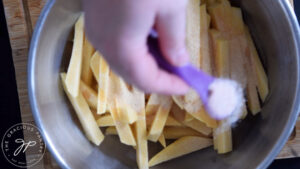 Adding salt to raw rutabaga fries in a mixing bowl.