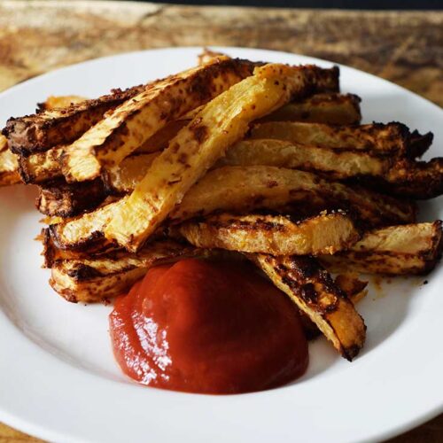 A white plate sitting on a wooden surface holds a pile of Air Fryer Rutabaga Fries with a dollop of ketchup.