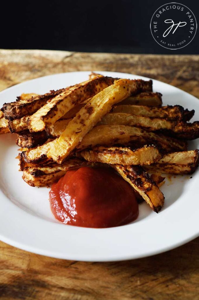 A white plate sitting on a wooden surface holds a pile of Air Fryer Rutabaga Fries with a dollop of ketchup.