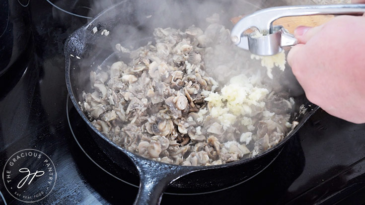 Adding pressed garlic to a skillet of cooked mushrooms.
