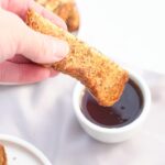 A female hand dunks a French Toast Stick into a small bowl of maple syrup.
