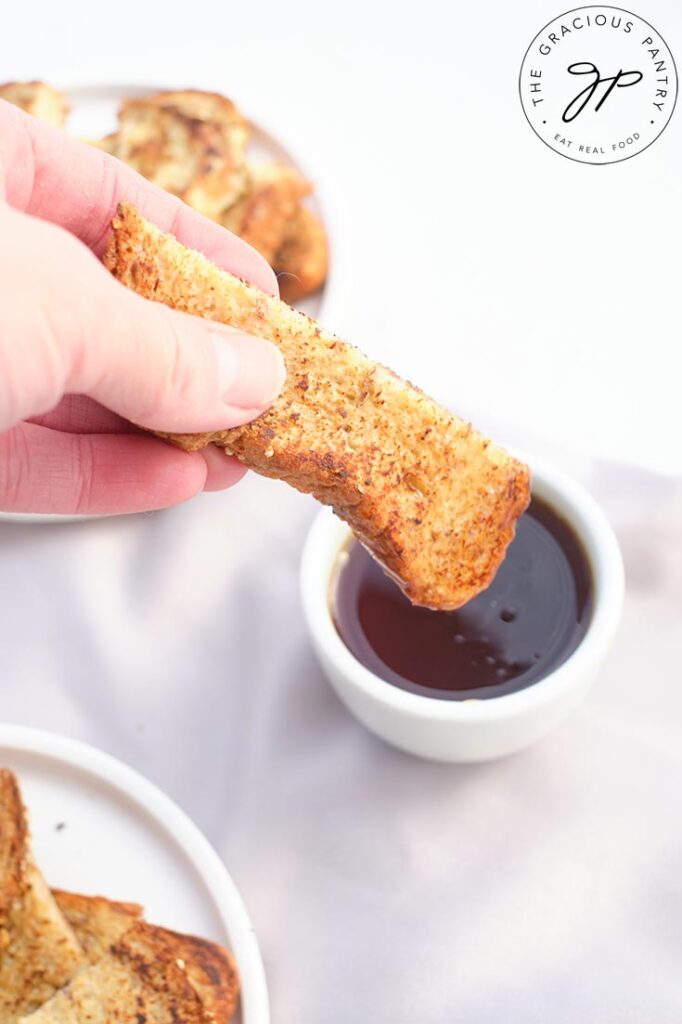 A female hand dunks a French Toast Stick into a small bowl of maple syrup.
