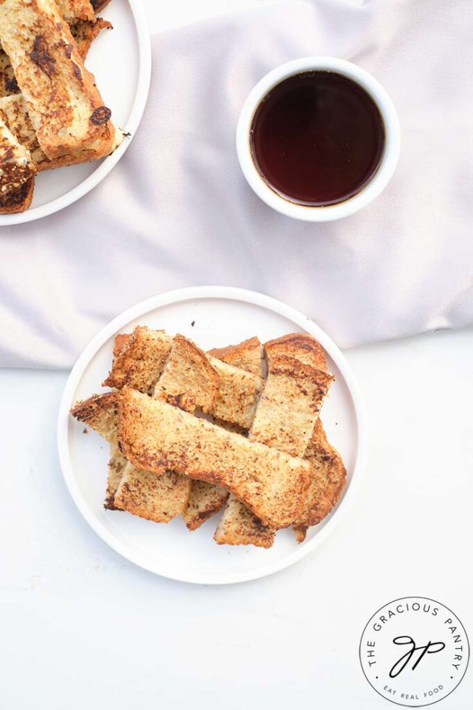 A white plate piled high with French Toast Sticks. A bowl of maple syrup sits next to it.
