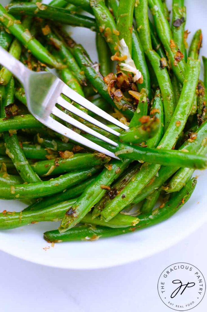 A closeup of Garlicky Green Beans in a white dish with a fork lifting up two green beans.