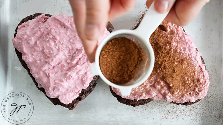 Sprinkling unsweetened cocoa powder over two frosted Healthy Chocolate Raspberry Cakes.