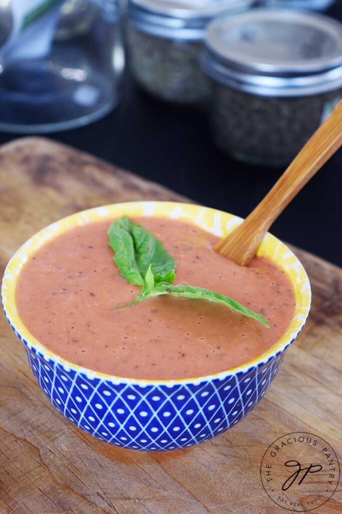 A blue and yellow bowl holds Healthy Tomato Soup and a wooden spoon. The soup is garnished with whole, fresh basil leaves.