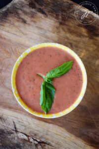 An overhead view of a bowl filled with Healthy Tomato Soup and garnished with whole, fresh basil leaves.
