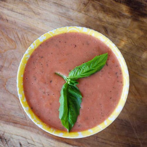 An overhead view of a bowl filled with Healthy Tomato Soup and garnished with whole, fresh basil leaves.