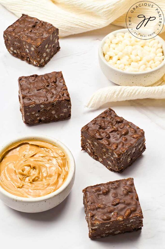 Homemade Crunch Bars on a white surface with a bowl of puffed brown rice cereal and a bowl of peanut butter.