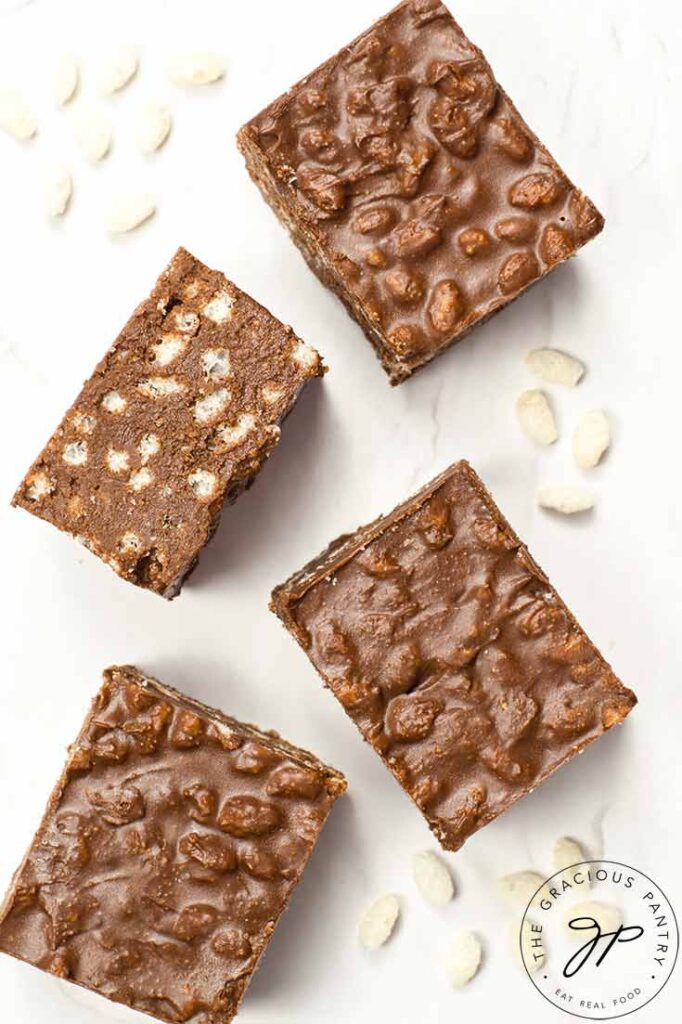 An overhead view looking down on four Homemade Crunch Bars and some puffed brown rice on a white surface.