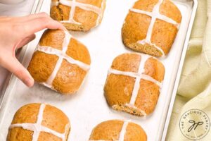 A female hand picks up a finished hot cross bun off of a baking pan.