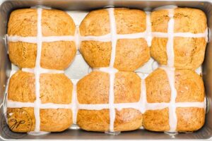 Hot cross buns sit in their baking pan with fresh icing crossed spooned over the top of each bun.