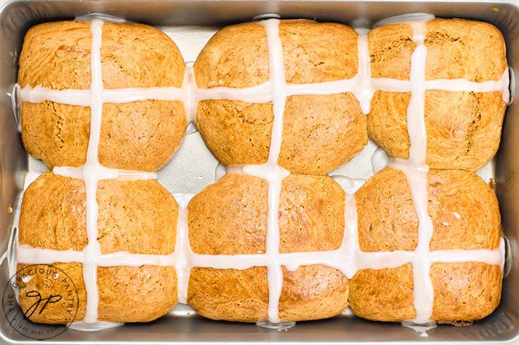 Hot cross buns sit in their baking pan with fresh icing crossed spooned over the top of each bun.