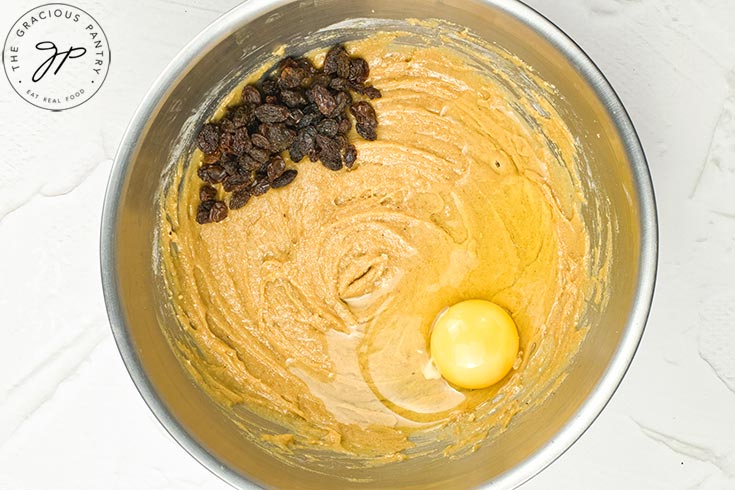 An egg and a small pile of raisins sits on raw hot cross bun batter, waiting to be mixed in.