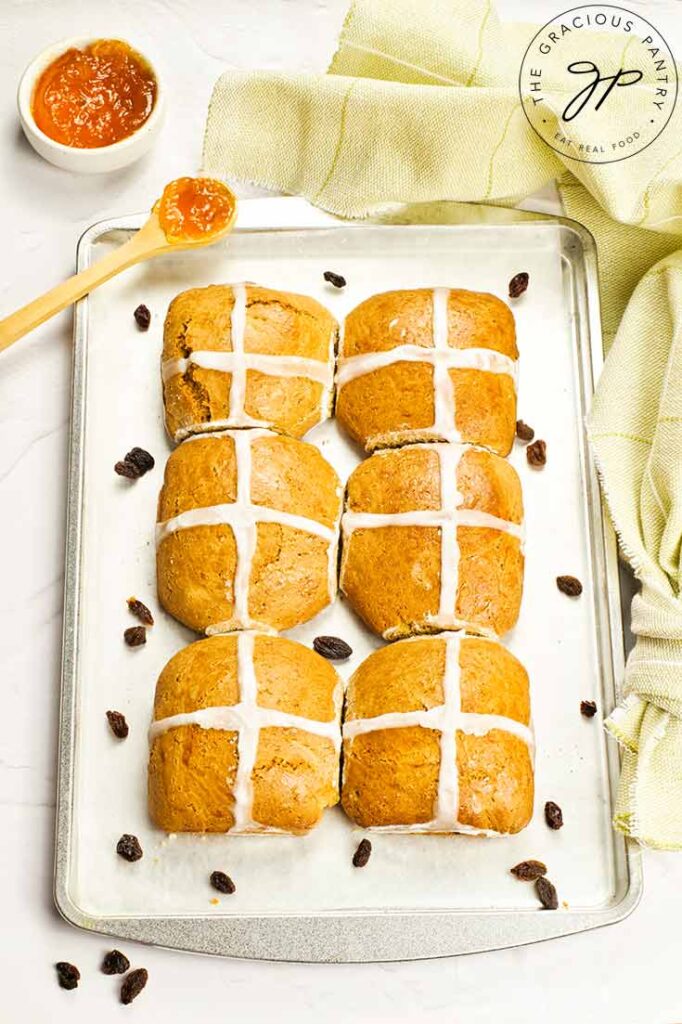 Six hot cross buns sit lined up on a baking pan with raisins sprinkled around them.