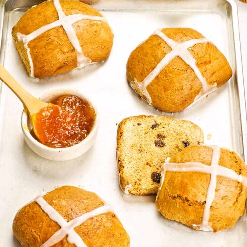 Four Healthy Hot Cross Buns sit on a baking pan with a bowl of marmalade.