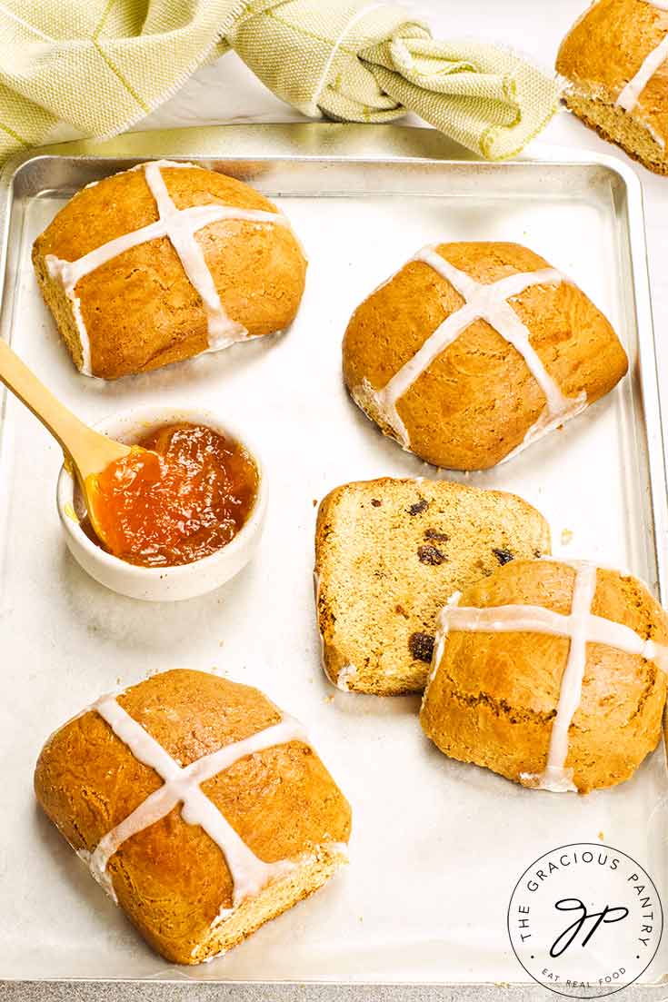 Four Healthy Hot Cross Buns sit on a baking pan with a bowl of marmalade.