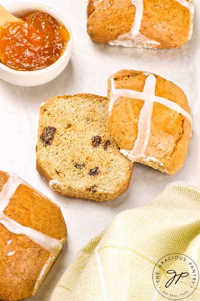 A cut Healthy Hot Cross Bun sits on a white surface next to two whole buns and a small bowl of marmalade.