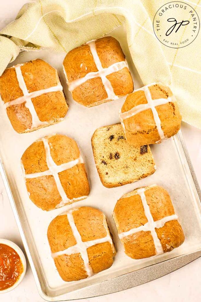 Healthy Hot Cross Buns sit on a baking pan lined up with one bun cut. The top rests to the side of the bun.