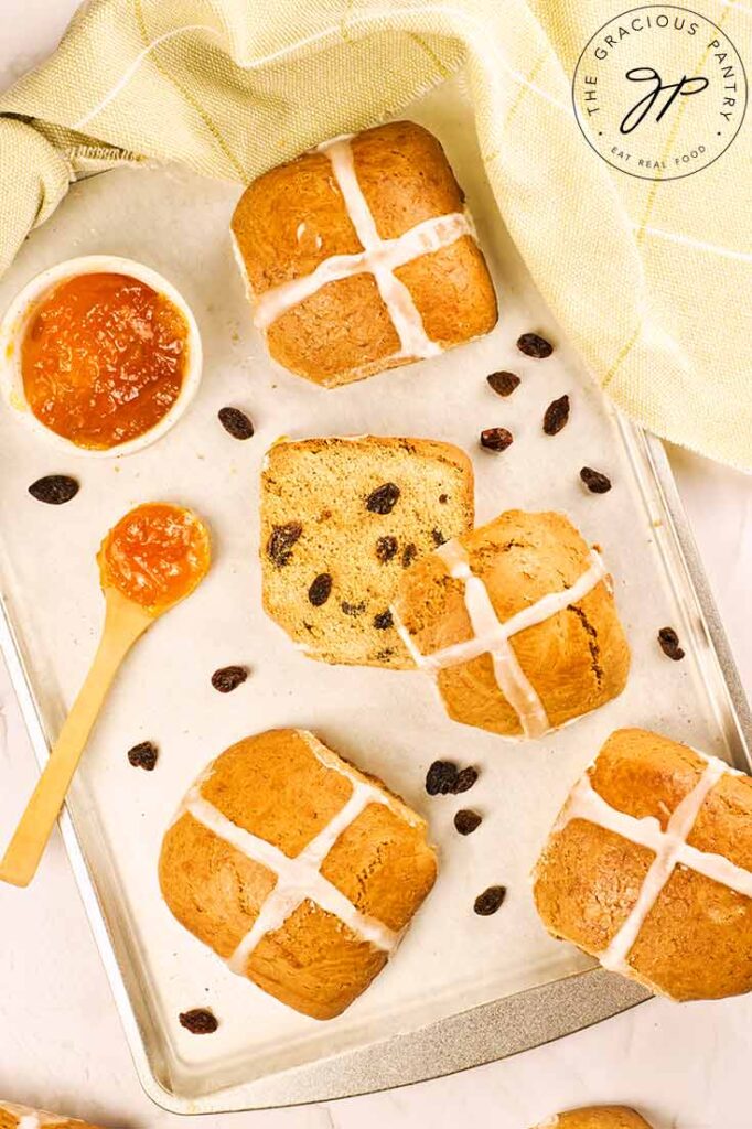 An overhead view of hot cross buns on a baking pan with raisins strewn around them and a bowl of jam sits to the side.