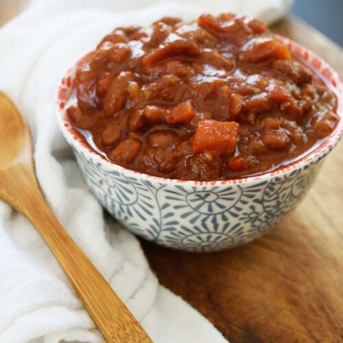 A side view of a bowl of Ranch Style Beans. A white towel and wooden spoon lay to the left of the bowl.