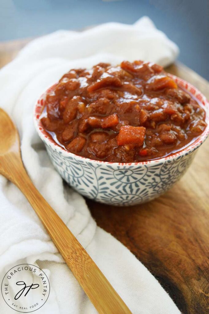 A side view of a bowl of Ranch Style Beans. A white towel and wooden spoon lay to the left of the bowl.