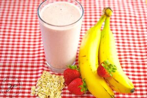 A red and white checkered tablecloth lays under a gathering of bananas, strawberries and oats with a glass of Strawberry Oatmeal Smoothie in their midst.