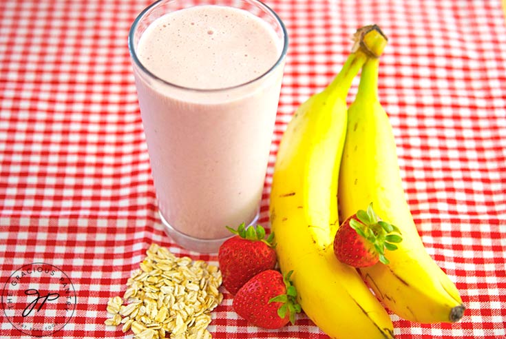 A red and white checkered tablecloth lays under a gathering of bananas, strawberries and oats with a glass of Strawberry Oatmeal Smoothie in their midst.