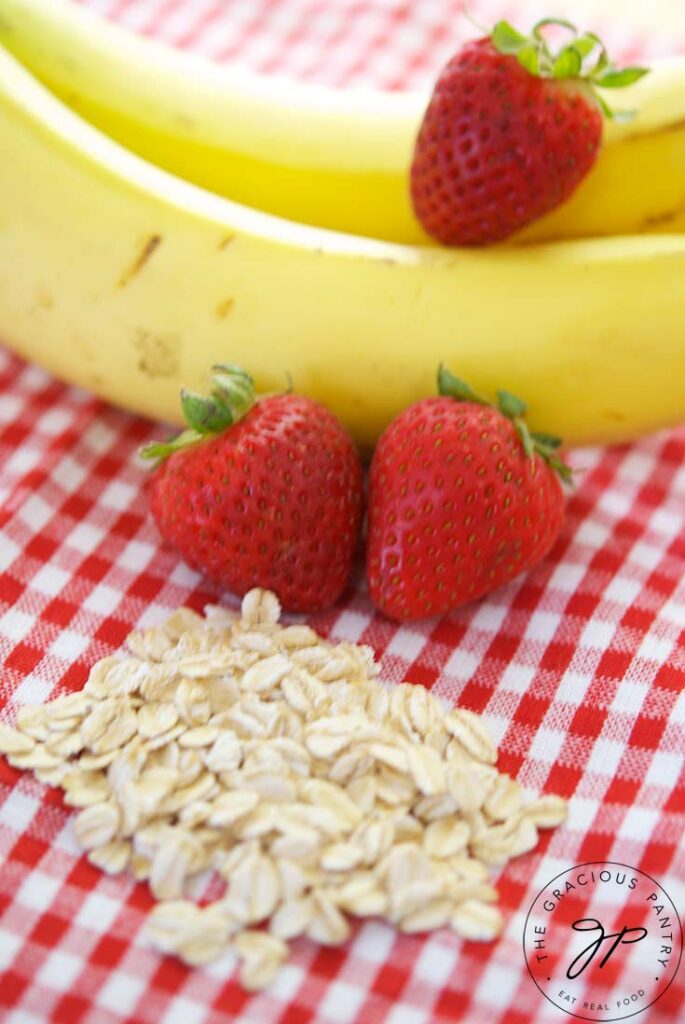 Two bananas, three strawberries and a small pile of oats gathered on a red and white checkered tablecloth.