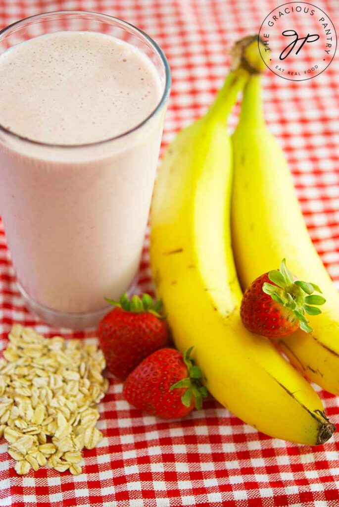 Fresh bananas, starwberries and oats lay gathered next to a glass of Strawberry Oatmeal Smoothie.