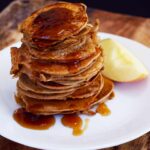 A stack of Healthy Apple Ring Pancakes with maple syrup over the top and sides, on a white plate.