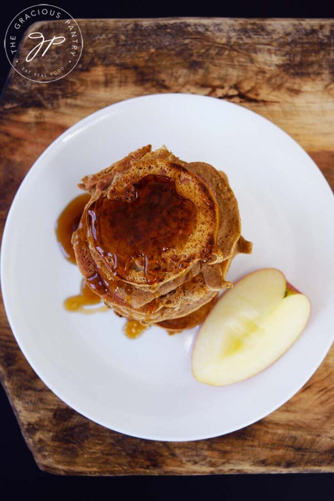 An overhead view of Healthy Apple Ring Pancakes topped with maple syrup. A piece of apple sits on the plate next to them.