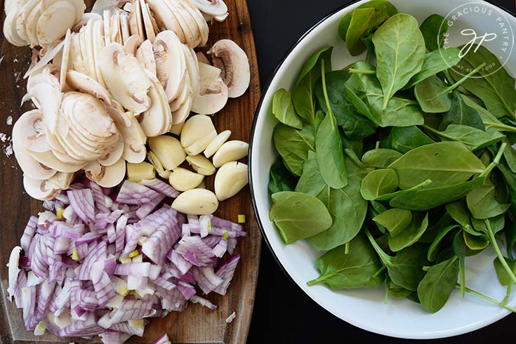 Chopped and sliced onions and mushrooms sit on a cutting board next to a bowl of raw spinach leaves.
