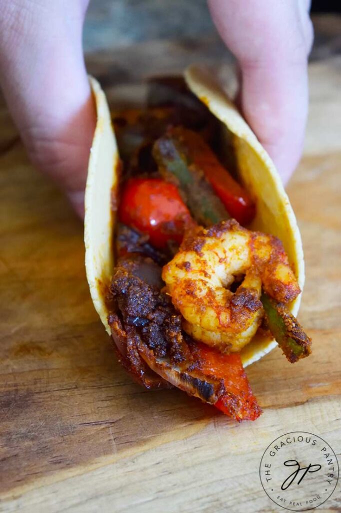 A female hand holds closed a single Sheet Pan Shrimp Fajita. A shrimp is at the front.