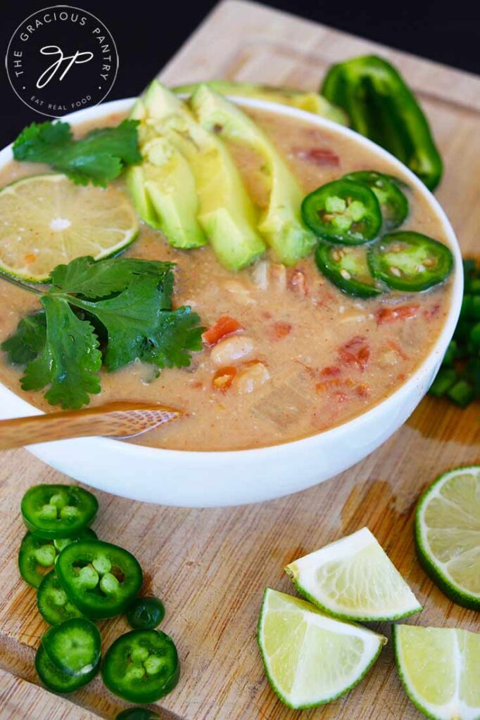 A side view of a white bowl filled with Crockpot White Chicken Chili and garnished with lime, cilantro, avocado and jalapeno peppers.