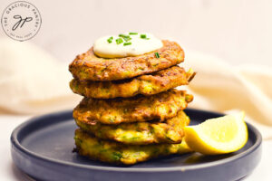A stack of finished Zucchini Fritters sit on a plate with a lemon wedge. The top fitter has a dollop of sauce on top of it.