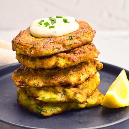 A tall stack of Zucchini Fritters sit on a plate with a lemon wedge, garnished with a dollop of sauce.