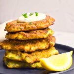 A tall stack of Zucchini Fritters sit on a gray plate with a wedge of lemmon.